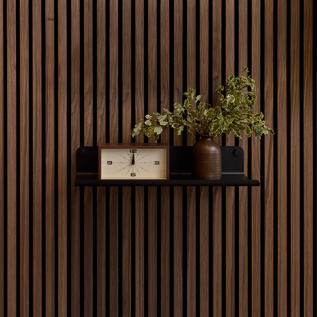 Black wood slat wall shelf mounted on vertical wooden slat wall, displaying a clock and potted plant.