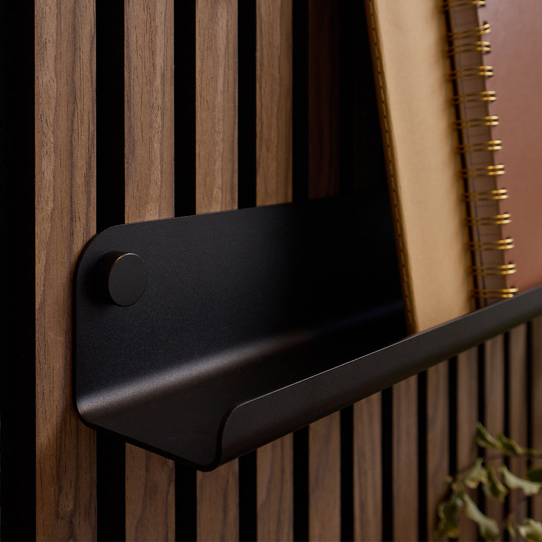 A matte black metal wall-mounted shelf bracket holding a notebook against a dark wood slat wall.