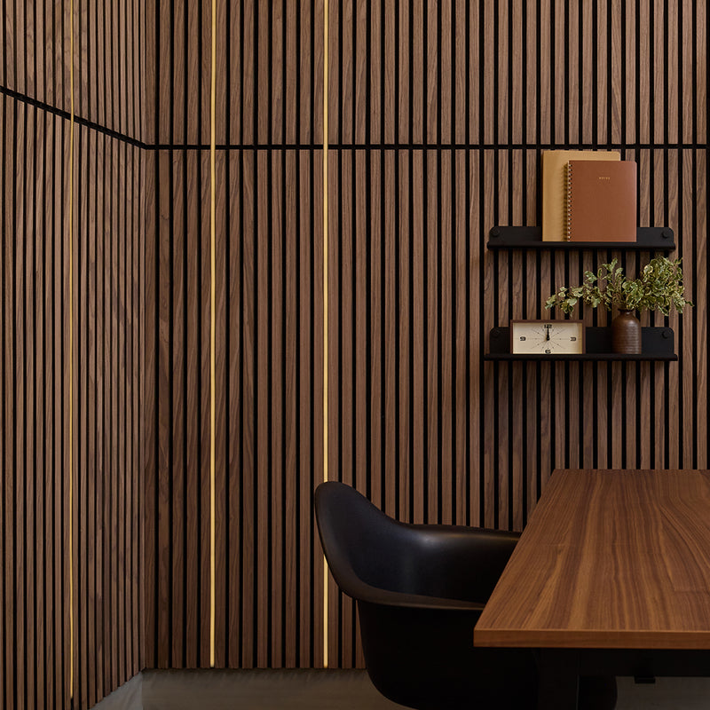 A modern interior scene with a dark wood slatted wall, black floating shelves holding books and small plants, a wooden desk, and a black chair in the foreground.