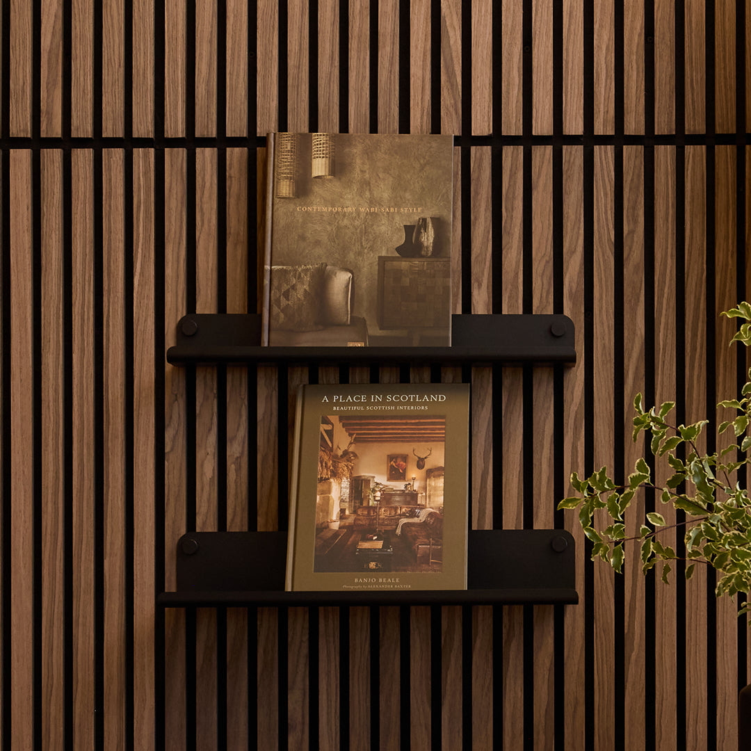 Two hardcover books displayed on black wall-mounted shelves against a dark brown wood slat wall, with a small green plant visible at the right edge.