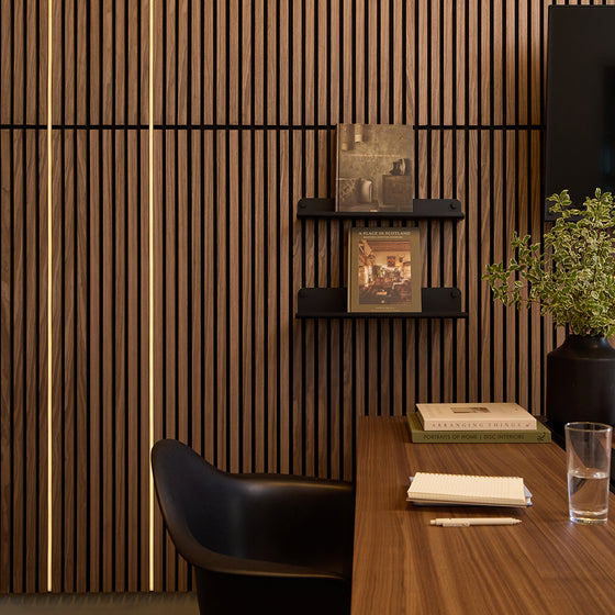 A modern interior scene with a dark wood slatted wall, black floating shelves, a black chair, a wooden desk, books, a pen, a glass of water, and a potted plant.