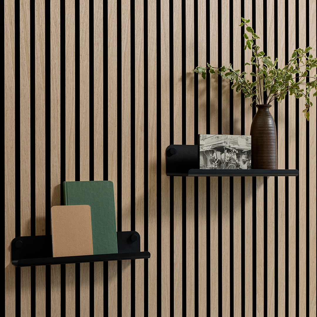 Two black floating wall shelves mounted on a light wood slatted wall, styled with books, a framed print, and a brown vase with greenery.