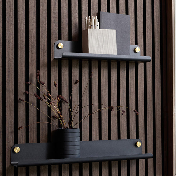 Two matte black wall-mounted shelves with gold-toned hardware, displayed against a dark brown wood slat wall. The top shelf holds a pencil holder and notebook, while the bottom shelf holds a black vase with dried stems.