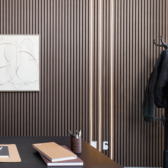 A modern interior scene with dark brown vertically slatted wall panels, a black desk, stacked notebooks, a pen cup, a framed abstract print, and a coat rack holding a dark jacket.