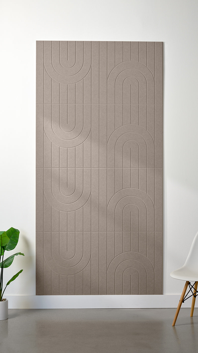 Greyish beige Felt Right 4’ x 8’ PET felt panel with a linear and curvy pattern engraved into the material against a white wall. A plant and chair are in the foreground.