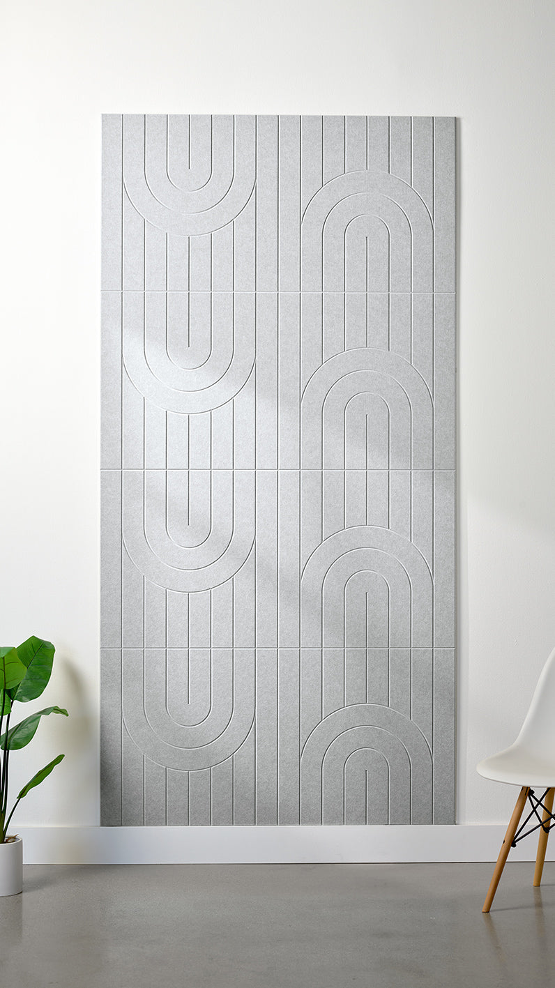 Heathered light grey Felt Right 4’ x 8’ PET felt panel with a linear and curvy pattern engraved into the material against a white wall. A plant and chair are in the foreground.