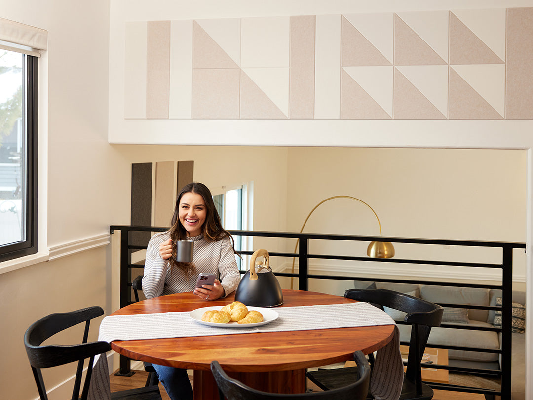 A Felt Right wall art design in cream shades on a wall above a kitchen table using triangles, squares and long rectangles. Woman drinking from a mug at the table