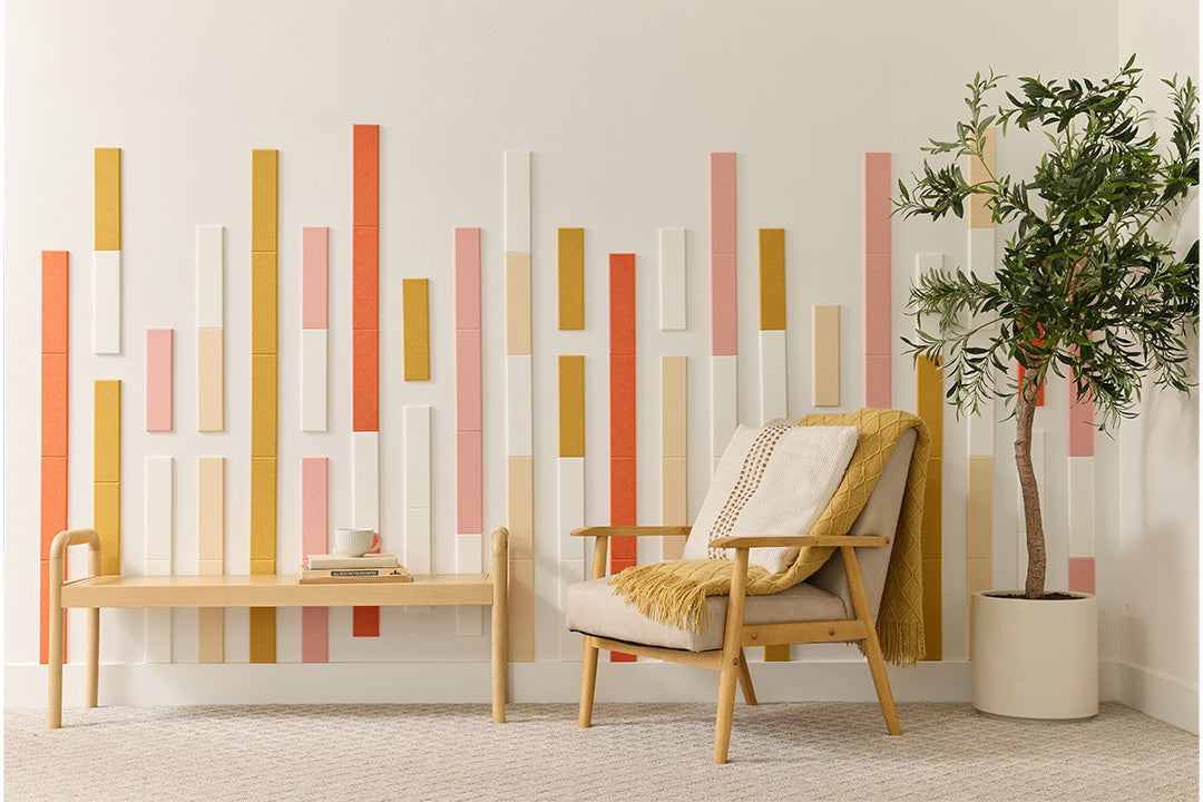 A room with a minimalist design featuring vertical bamboo peach panels in shades of beige, brown, and white arranged on a wall. There is a wooden bench, a white chair with a beige cushion, and a potted plant to the right.
