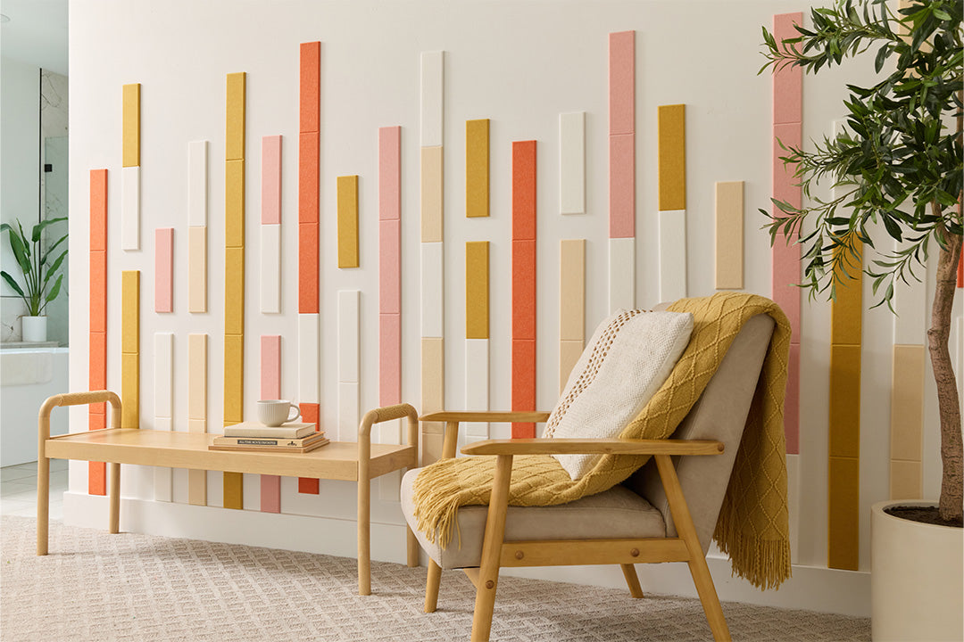 A room with a minimalist design featuring vertical bamboo peach panels in shades of beige, brown, and white arranged on a wall. There is a wooden bench, a white chair with a beige cushion, and a potted plant.