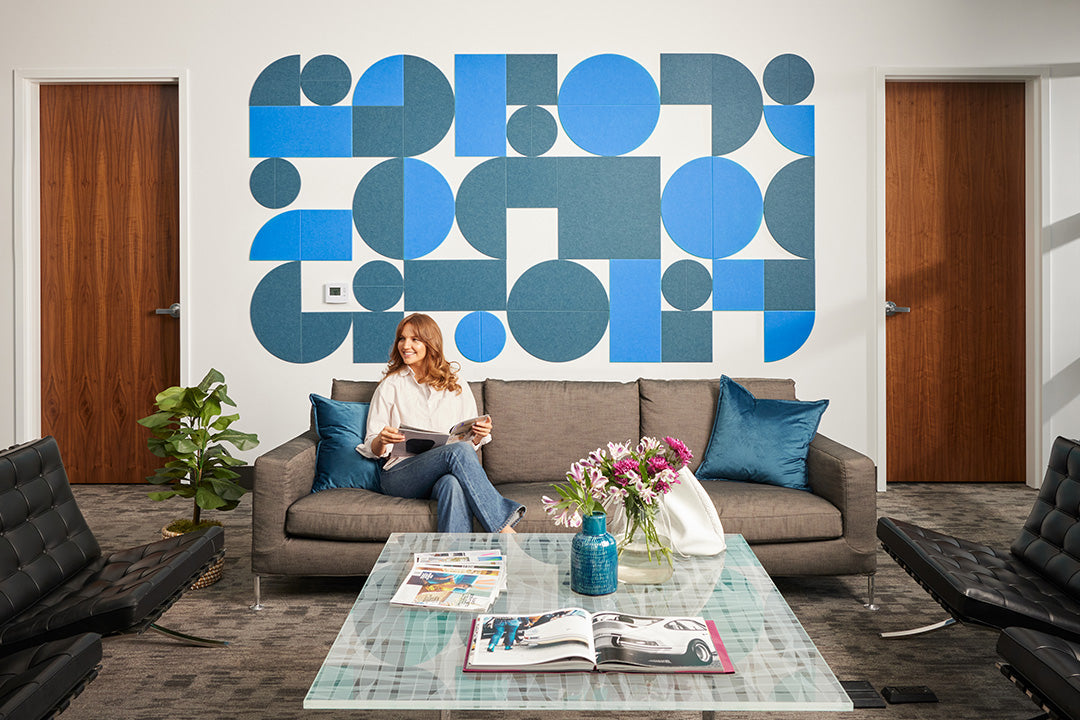 A Felt Right wall art design in shades of blue using circles and squares to create geometric design on an office wall with a couch and a coffee table and a woman sitting on the couch.