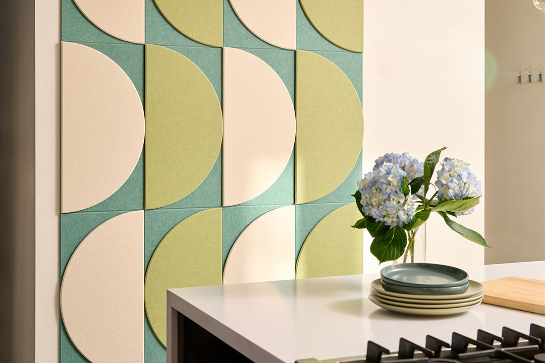 A Felt Right layered wall art design in shades of green and cream on a wall next to a counter with flowers.