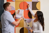 Man and woman installing the Layered Groovy Tuesday Felt Right design. The man is holding a paper with the instructions and the woman is installing an orange tile against a colorful felt background while holding a level.