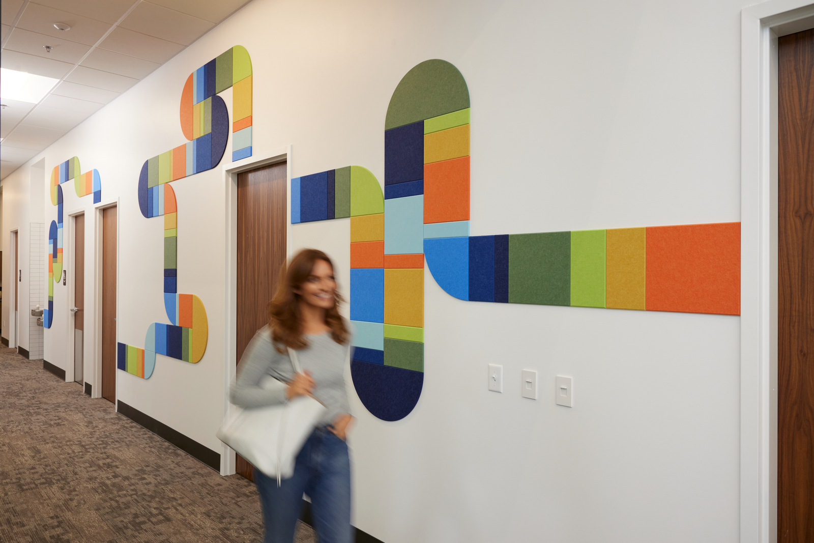 felt right wall art design in multicolors creating a pipe-like design going down a hallway with doors and a woman walking