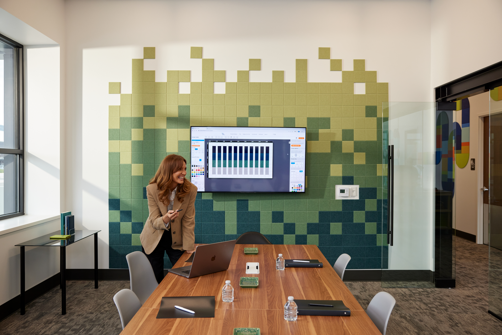 A Felt Right wall design in a conference room along the wall in a pixelated design in shades of green around a TV and a lady leading a discussion.