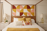 A Felt Right design wall in a bedroom setting with a bed featuring a geometric pattern headboard in shades of yellow, orange, and pink, accompanied by matching pillows and a quilt, with bedside tables and lamps.