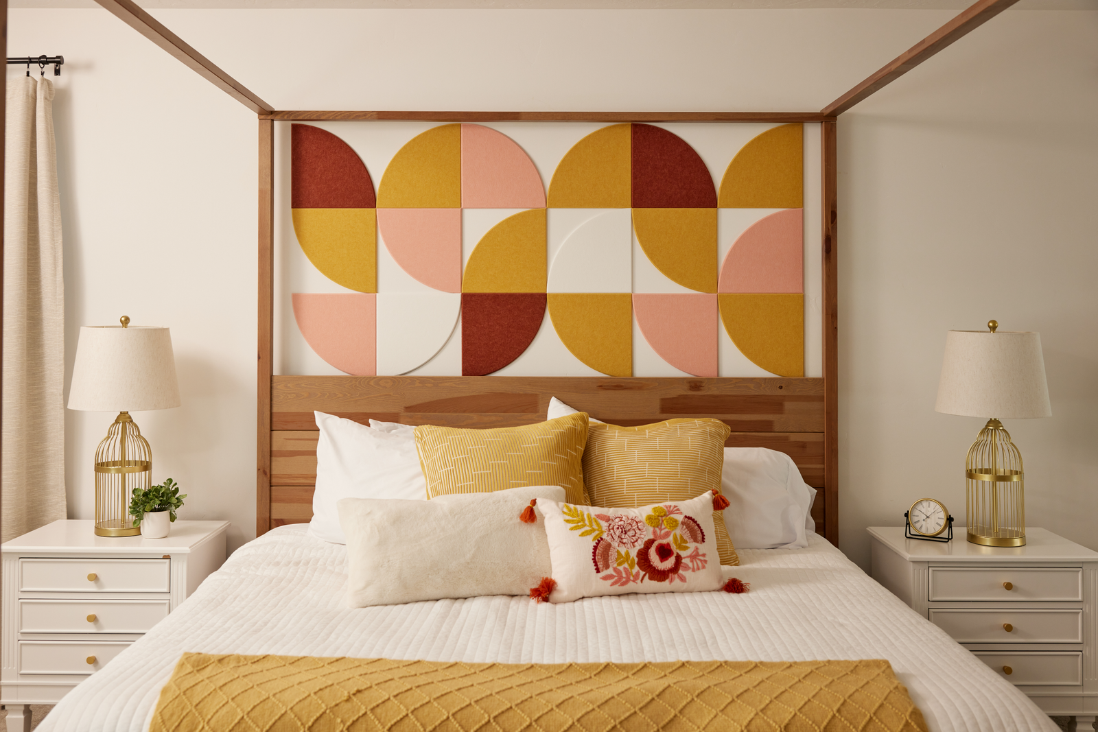 A Felt Right design wall in a bedroom setting with a bed featuring a geometric pattern headboard in shades of yellow, orange, and pink, accompanied by matching pillows and a quilt, with bedside tables and lamps.