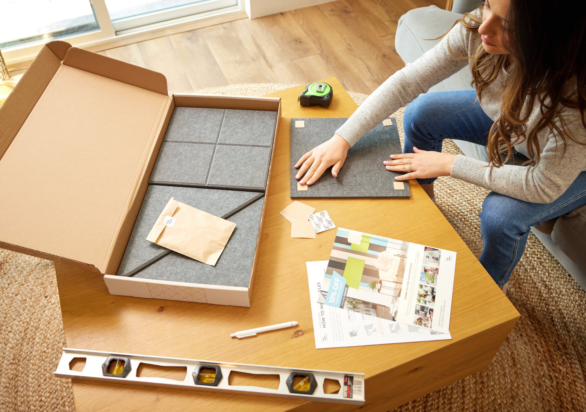 A woman is on her couch and placing Felt Right double-sided adhesive tabs on the back of her grey Felt Right tiles. There is a box of Felt Right tiles on the coffee table as well as a ruler, level, pen, and some papers about Felt Right.