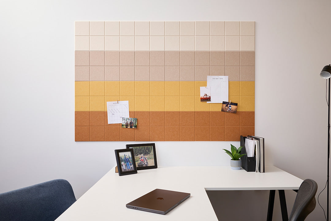 A Felt Right wall design bulletin board with a gradient of colors ranging from beige to ochre, mounted on a wall above a white desk with various items such as a photo frame, a potted plant, and a closed laptop.