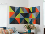 Felt Right wall art design that uses triangle felt in shades of green, grey, blue, and orange on a white walls in a corner of a room by a couch and a side table.