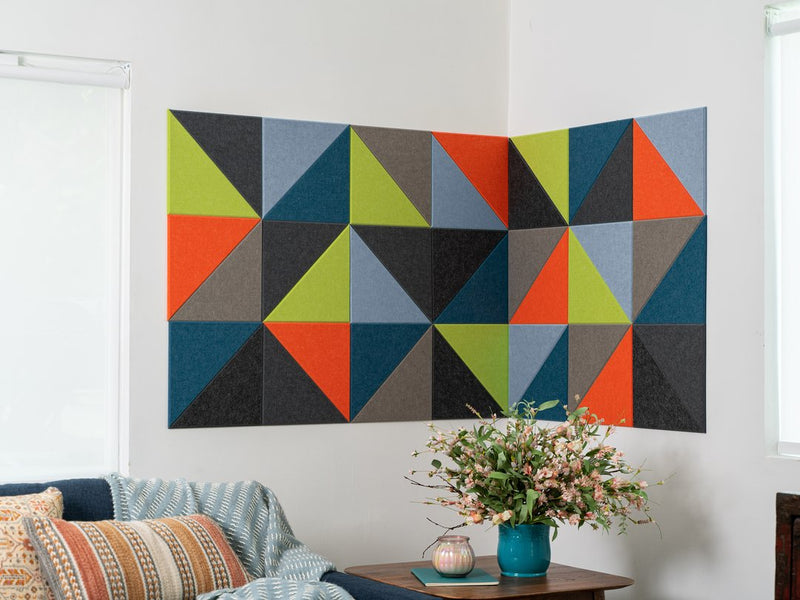 Felt Right wall art design that uses triangle felt in shades of green, grey, blue, and orange on a white walls in a corner of a room by a couch and a side table.