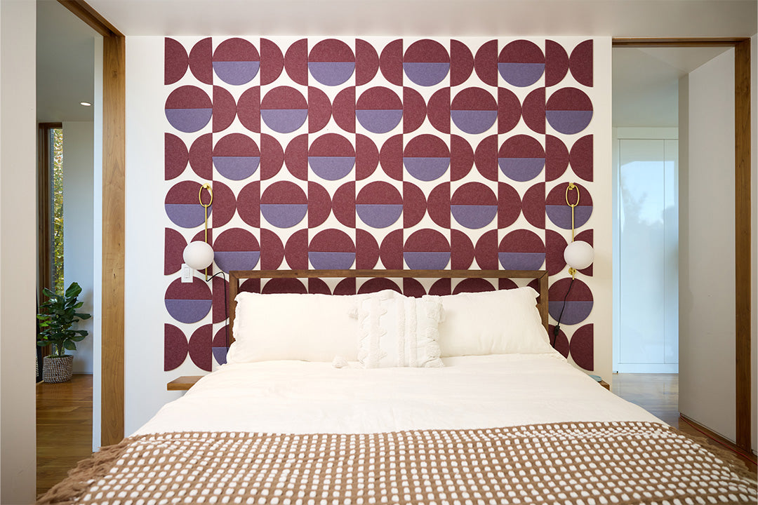 A Felt Right design using half circles in purple shades on a wall behind a bed with two lamps on the wall in a bedroom.