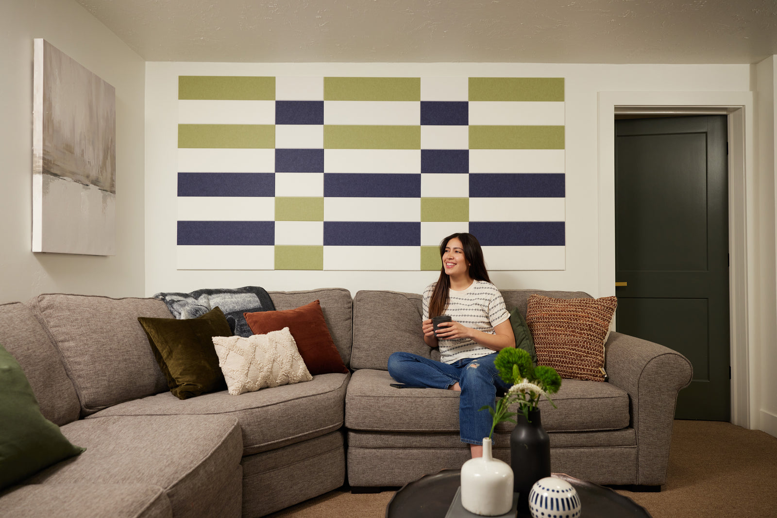 A Felt Right wall art design in shades of blue, green, and white on a white wall in a living room above a couch with a woman drinking from a mug