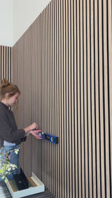 Woman installing black shelf on oak black wood slat wall paneling. 