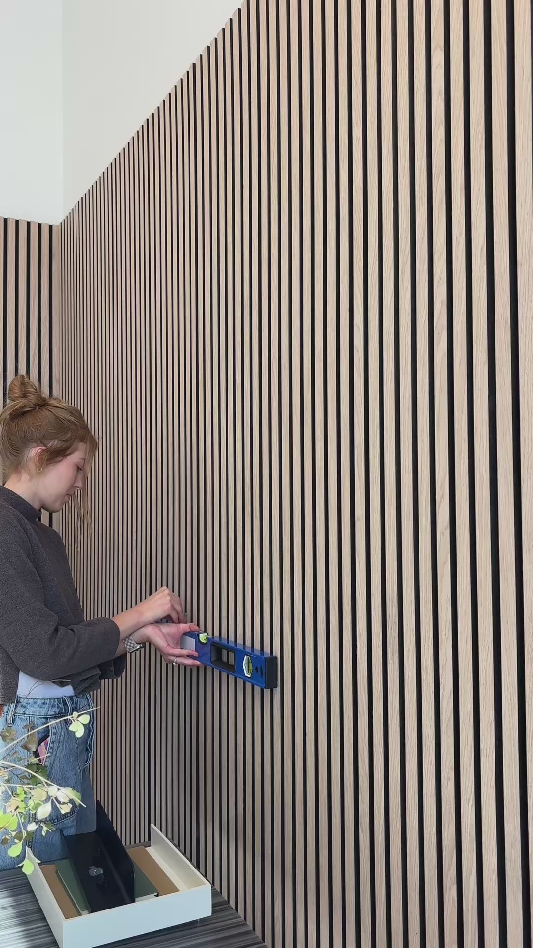 Woman installing black shelf on oak black wood slat wall paneling. 