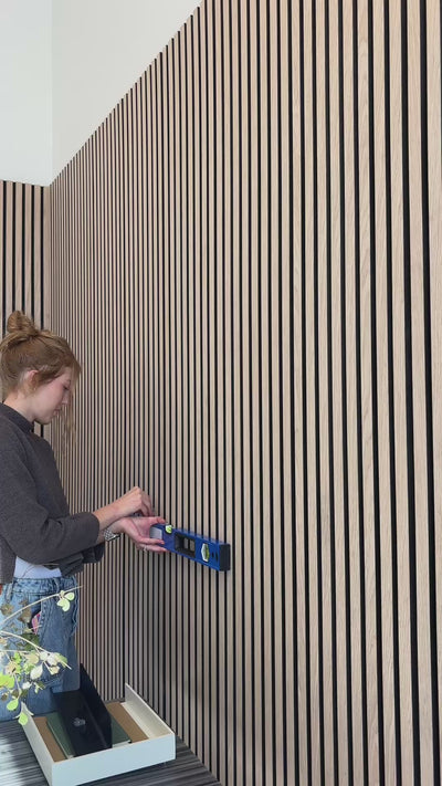 Woman installing black shelf on oak black wood slat wall paneling. 