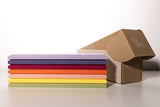 Stack of colored fabric samples in lavender, navy, red, orange, yellow, and green beside kraft cardboard box.