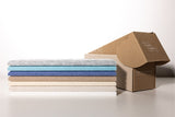 Stack of five folded fabric samples in gray, light blue, navy, tan, and cream beside an open kraft cardboard box.