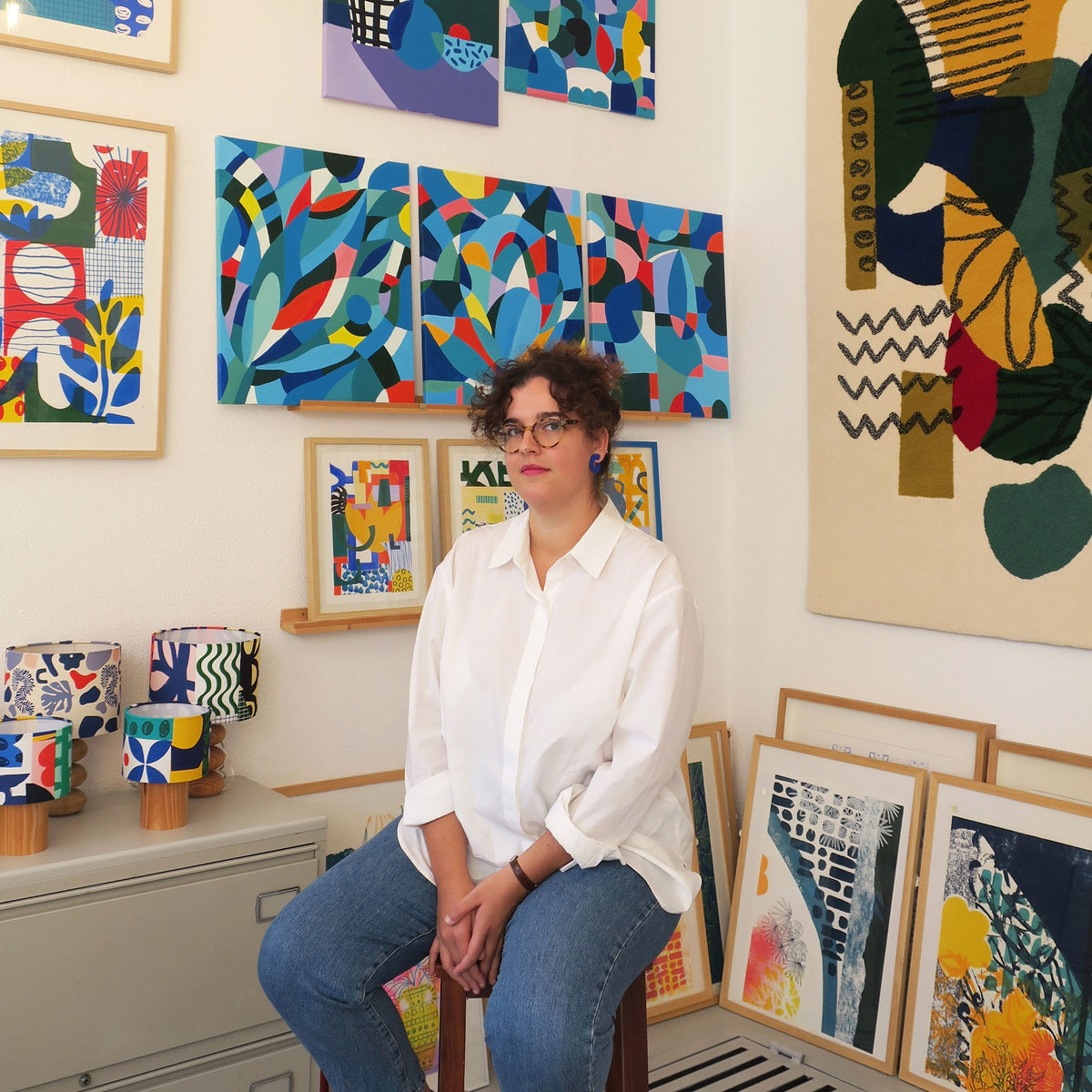 A woman with curly dark hair and glasses, wearing a white button-down shirt and blue jeans, sits in a room filled with colorful abstract art prints and patterned home decor items.