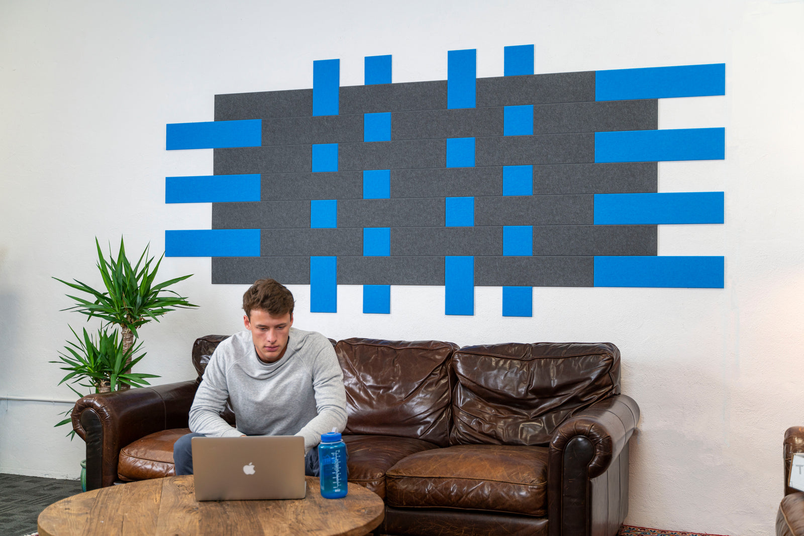 A Felt Right wall art design in shades of grey and blue felt tiles on a white wall behind a couch with a man working on his computer.