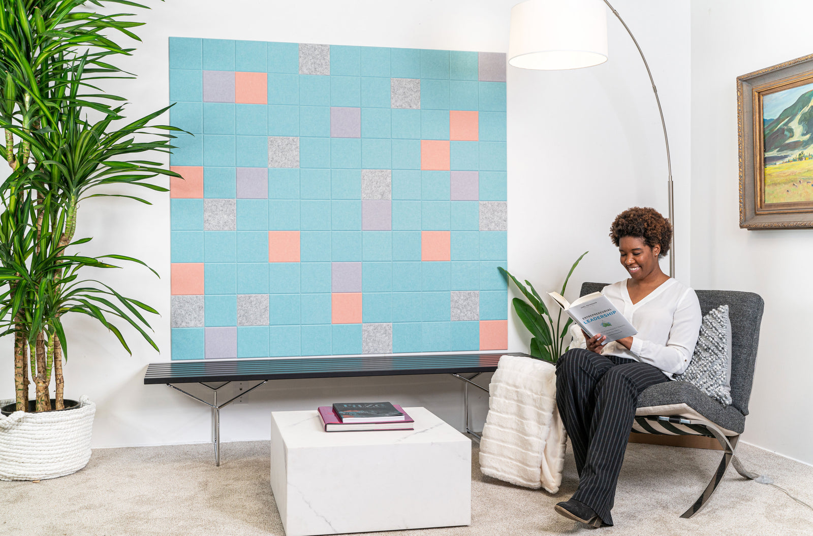 A Felt right wall art design in shades of blue, pink, purple, and grey felt tiles on a white wall in a waiting room with a woman in a chair reading a book.