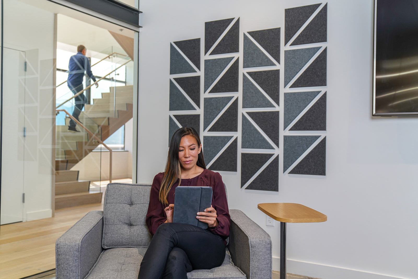 A Felt Right wall art design in shades of grey felt tiles on a wall in a room with a woman sitting in a chair on an iPad.