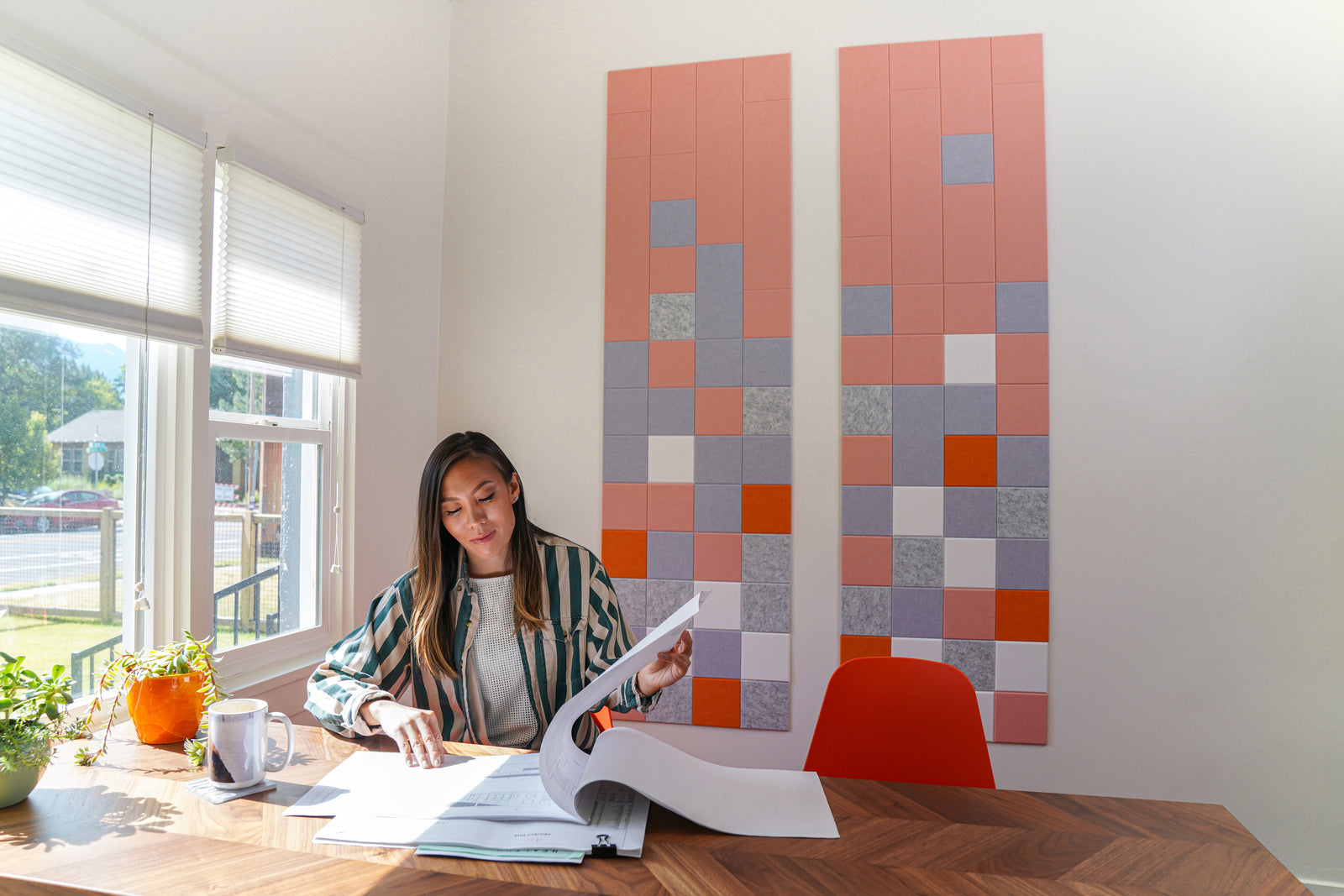 A Felt Right wall art design using shades of pink grey and white pixelated squares on a white wall in a dinning room or office with a woman working at the table.