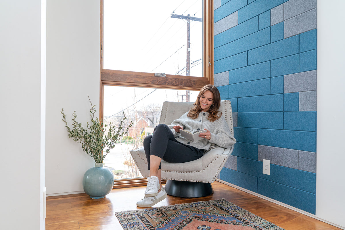 A Felt Right wall art design in shades of blue and grey felt tiles on a white wall with a woman reading in a chair.