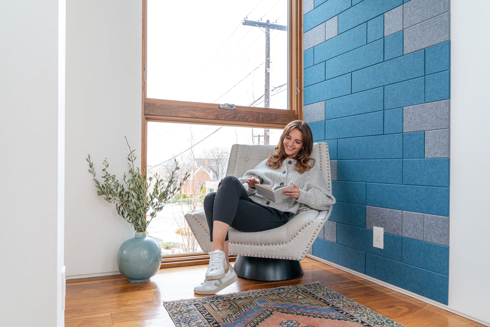 A Felt Right wall art design in shades of blue and grey felt tiles on a white wall with a woman reading in a chair.
