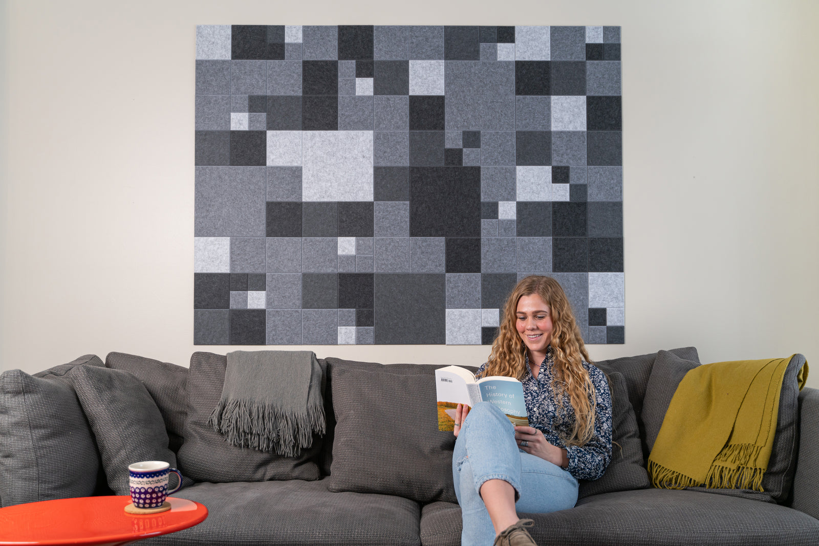 A Felt Right Wall art design in shades of grey felt tiles on a wall behind a couch with a woman reading on it.