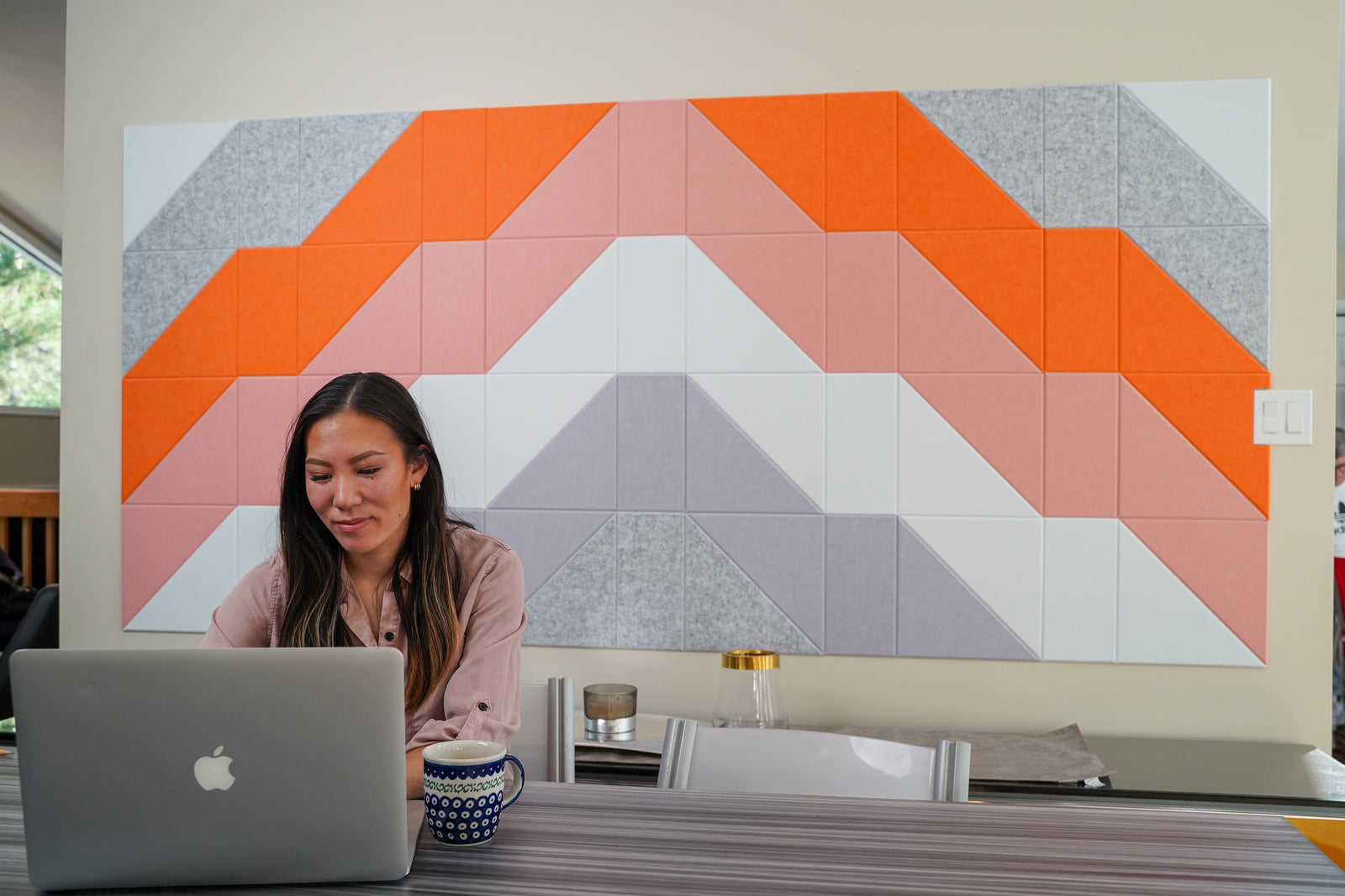 A Felt Right wall art design in shades of grey, orange, pink, and purple making a geometric summit pattern on felt tiles on a wall with a woman working at her desk.