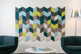 A Felt Right wall art design in shades of blue, green, and greys creating a geometric pattern on an white wall in between two chairs and a side lamp.