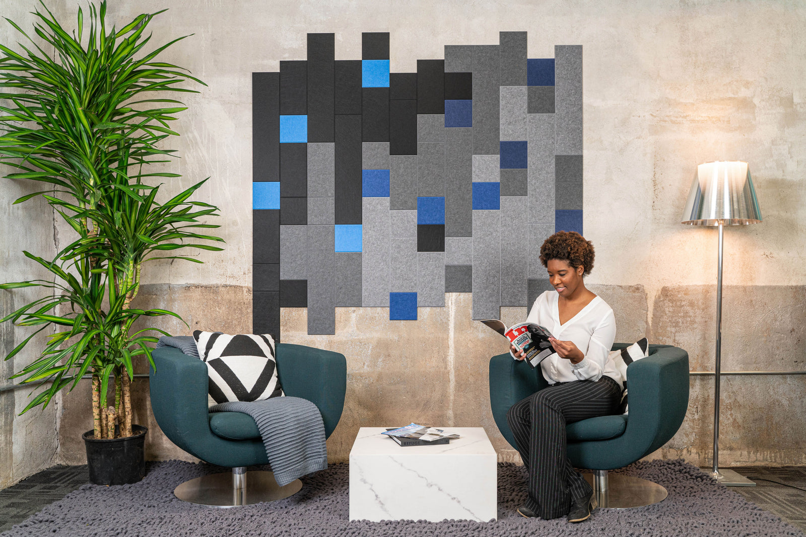 A Felt Right wall art design in shades of grey, black, and blue felt tiles on a wall with two chairs and side table in a waiting room with a woman reading a magazine.
