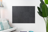 A Felt Right wall design with parallel lines in a woven look, on a white wall in a dark grey shade, acoustic, in a living space next to a couch and a plant.