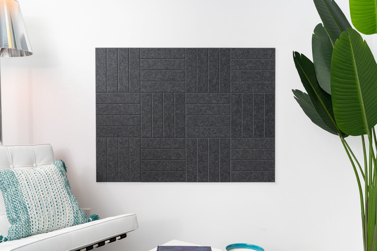A Felt Right wall design with parallel lines in a woven look, on a white wall in a dark grey shade, acoustic, in a living space next to a couch and a plant.