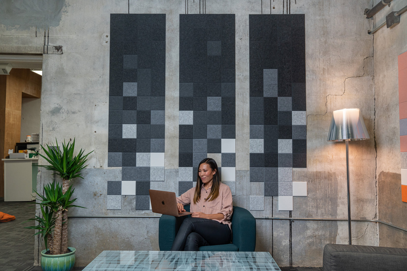 A Felt Right wall art design in shades of grey in a pixelated square design on a cement wall next to a lamp and a woman in a chair working on her laptop.