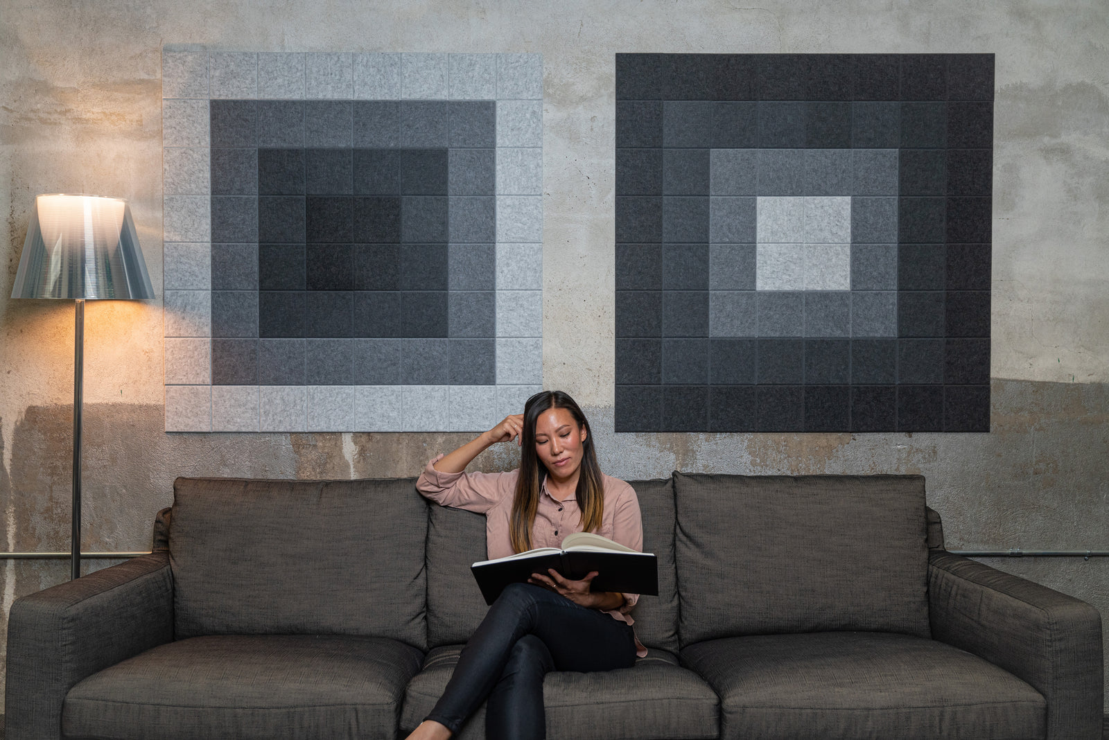 A Felt right wall art design in shades of grey square felt tiles on a cement wall  with a woman working on a couch.