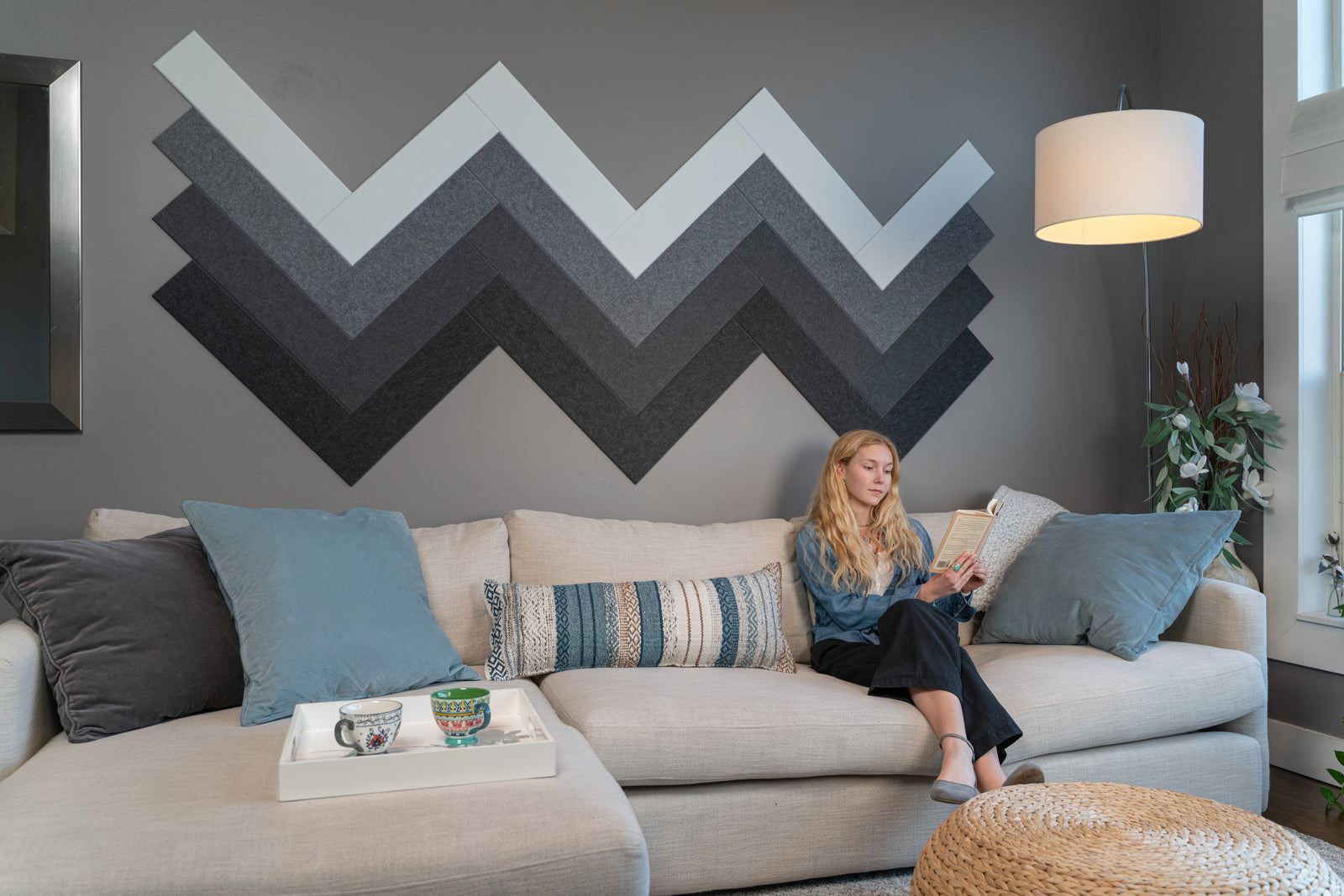 A Felt Right wall art design in shades of white, grey, and black in felt tiles making a zig zag pattern on a grey wall with a woman reading on the couch