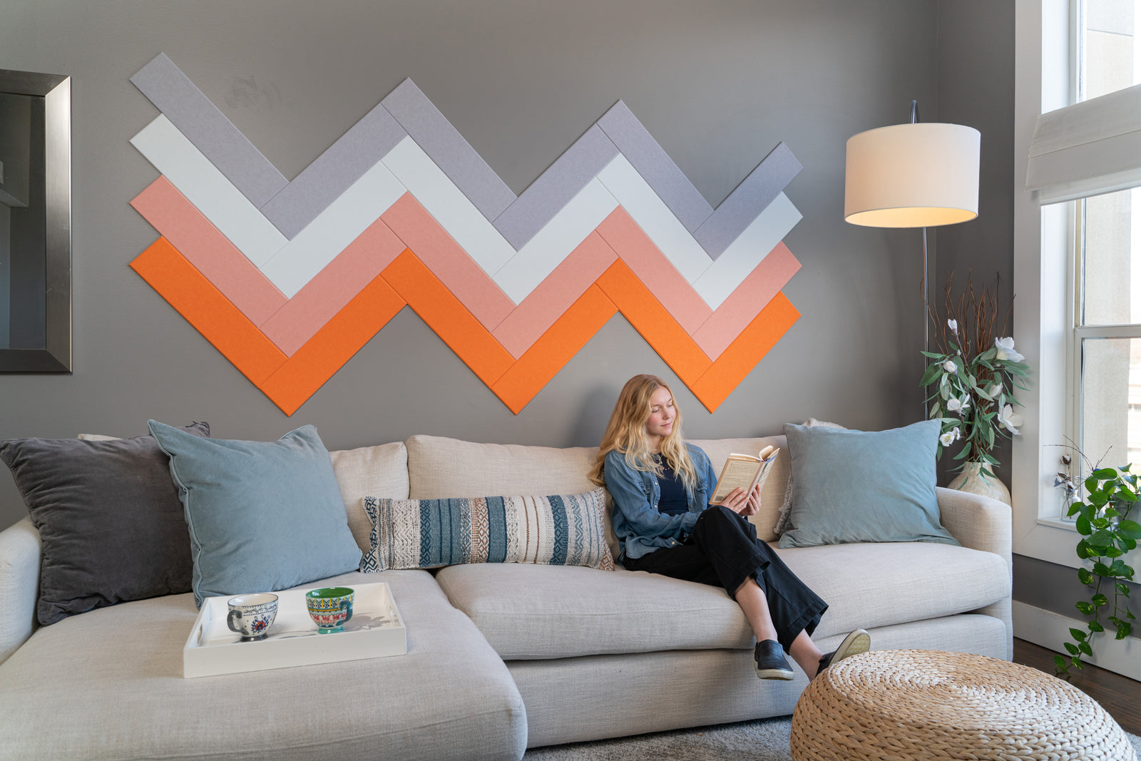 A Felt Right wall art design in shades of purple, white, pink, and orange on a grey wall with a woman reading on the couch.