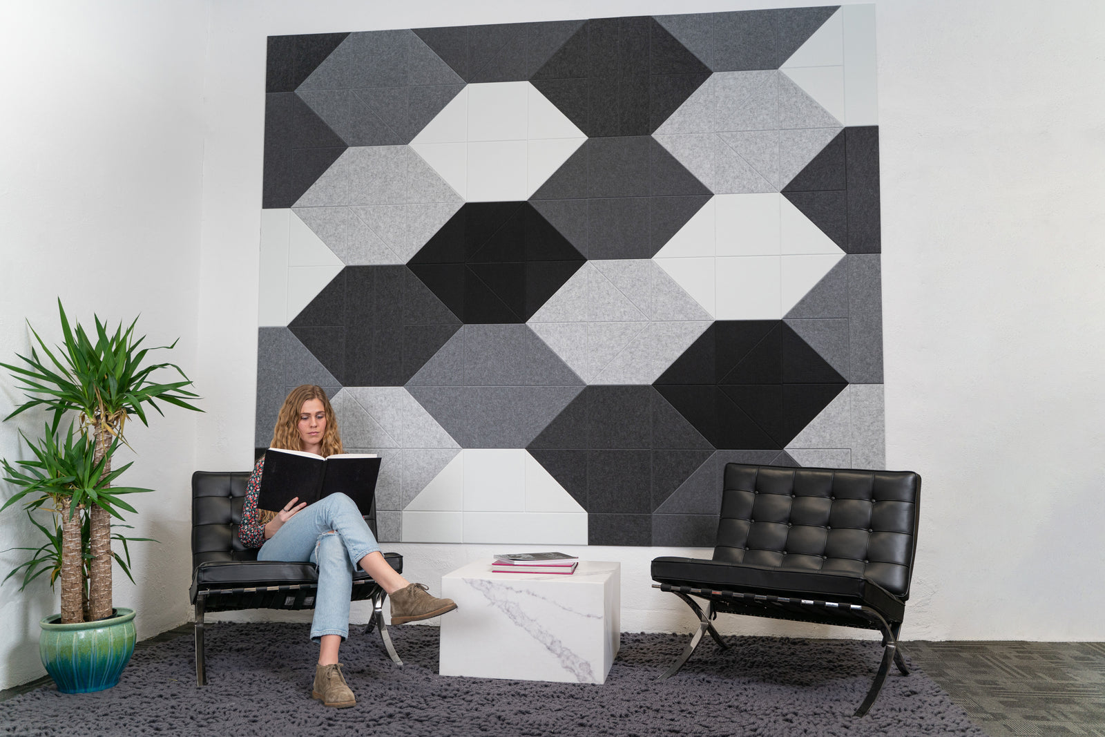 A Felt Right wall art design in shades of black and white felt tiles on a white wall with two chairs and a side table in an office or a waiting room.