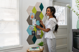 A Felt Right wall art design in shades of grey, green, blue, and cream creating a geometric 3d cubes that are pinnable next to a door with a women pinning cards on it.